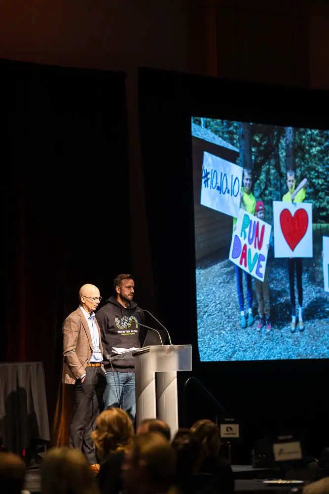 AbaData User Conference 2025 Images (12 of 20) showing a heartwarming photo of a supportive family with hand-coloured signs for their dad, as David Morin and Mike Hanson share from the podium about the impact of their 10-in-10-in-10 Marathon Challenge