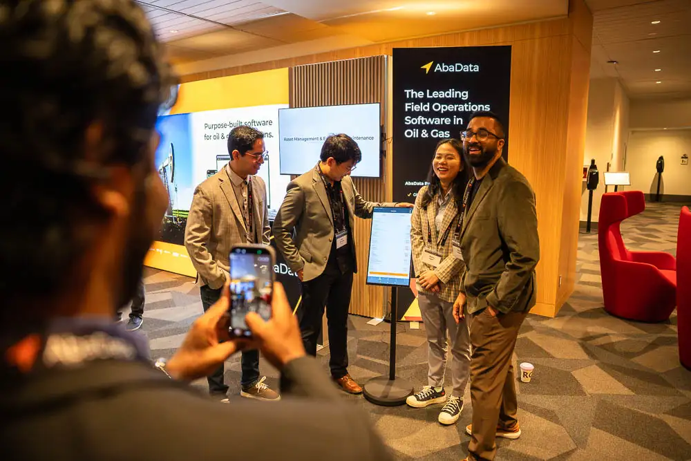 AbaData User Conference 2025 Images (2 of 20) showing joyful AbaData developers laughing together as they stand proudly in front of a large demo of the mobile software they have built
