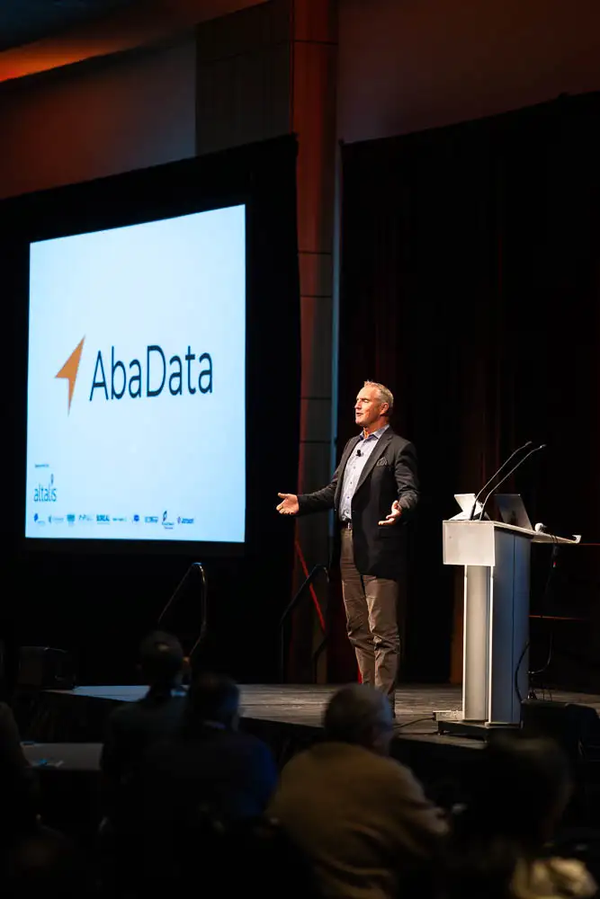 AbaData User Conference 2025 Images (5 of 20) showing keynote speaker Mark Little as he enthusiastically delivers his address with his arms spread wide with an AbaData logo on a screen behind him