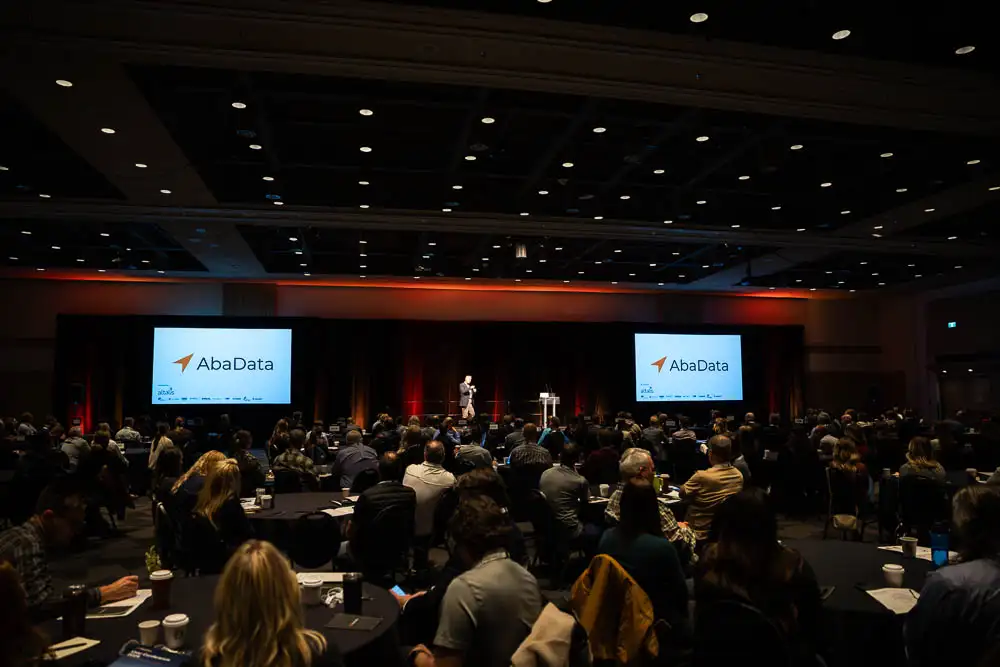 AbaData User Conference 2025 Images (6 of 20) showing a full room of attendees listening to an opening address from our keynote speaker Mark Little, with AbaData logos on two side screens